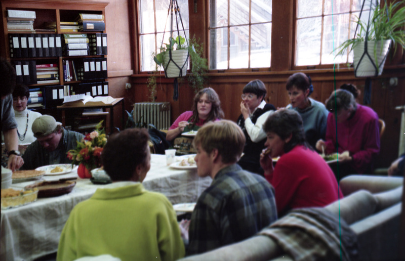 People sitting on couches in the Women's Center eating food. They surround a table filled with plates with food. Judy Wallins is in the center of the image, sitting against the wall with her hand to her mouth. Valerie Russo (in green) sits in the front next to Kelly Anderson (Jill Anderson's son).