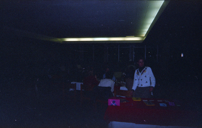 A dimly lit photo with people in it. A woman in a white sweater stands in front of a red table that has what appears to be fliers. Other people can be seen sitting at tables in the background.