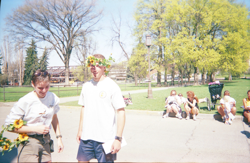 Two people hold flower crowns in the foreground while people sit on the curb in the background. Construction of the Idaho Student Union Building can be seen in the background.