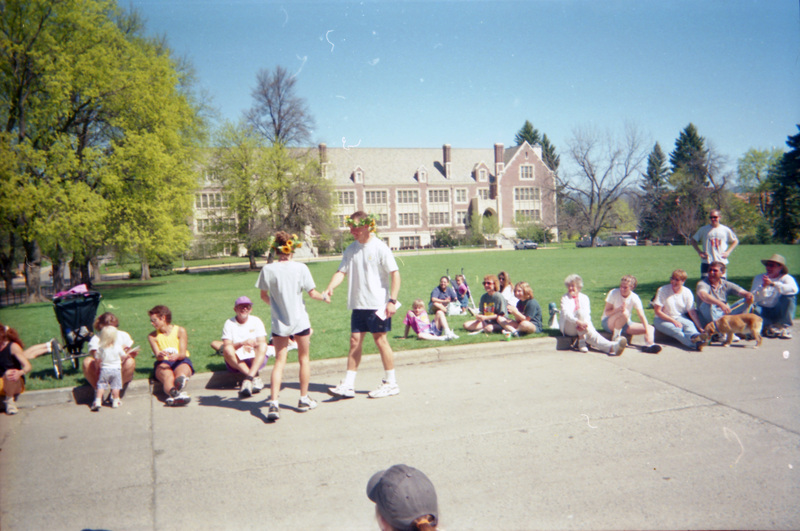 Two people wearing flower crowns go to shake hands, while other people sit on a curb watching. The Administration Lawn can be seen in the background.