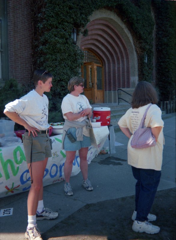 Three women stand near a table outside the Administration Building. A table with cups, a water color, and a hand-painted banner can be seen behind the women standing.