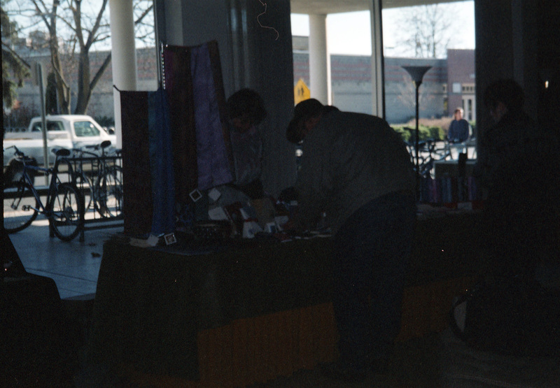 Three people stand near a booth inside a building. There are what looks like pieces of fabric displayed on the table.