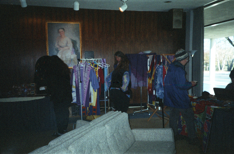 Three people looking at items; one person is looking at a clothing rack while another is speaking to someone at a booth.