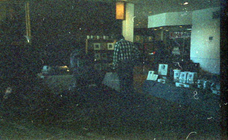 Several people looking at items at booths. The tables are covered with cloth and filled with various items including framed images and pottery. The image is faded and blurry.