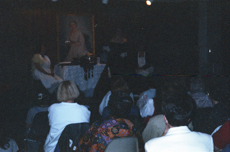 People sitting in an audience and watching a speaker dressed in a long black gown. There is another woman sitting near the speaker and a painted portrait in the background.