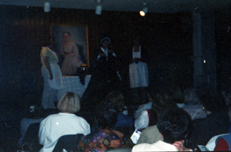 Three women in older clothing (aprons, bonnets) presenting something to a seated audience. 