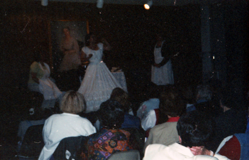 Three women wearing older clothing presenting something to a seated audience. A woman wearing a long white dress stands in the center.