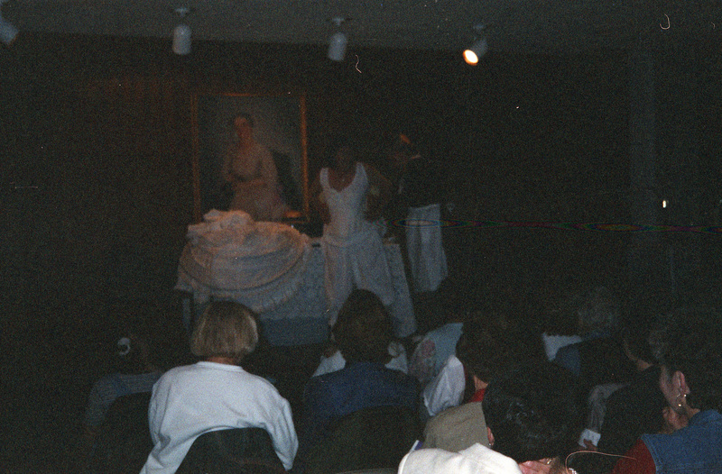 Two women stand before an audience. They are both wearing older clothing. A white piece of clothing is on the table next to them.