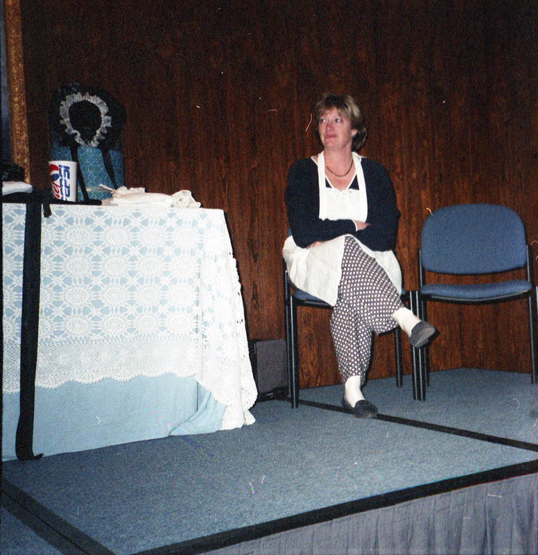 Jill Anderson wearing an apron sits on a stage next to a table with objects on it. There appears to be what looks like a hatbox on the table.