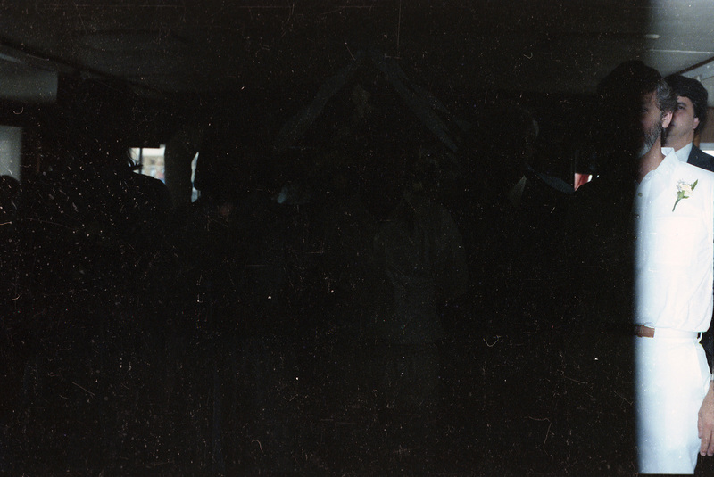 A dimly lit photograph of people posing. Two men can be partially seen on the right, one of which is wearing all white and has a flower pinned to their shirt.