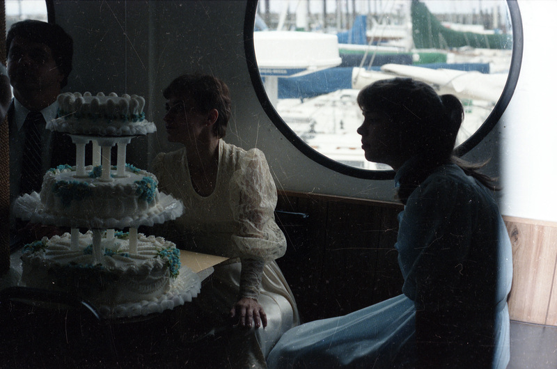 A bride and groom sitting behind a wedding cake. A young girl in a dress sits beside them. The circular window behind all three people show a view of a harbor.