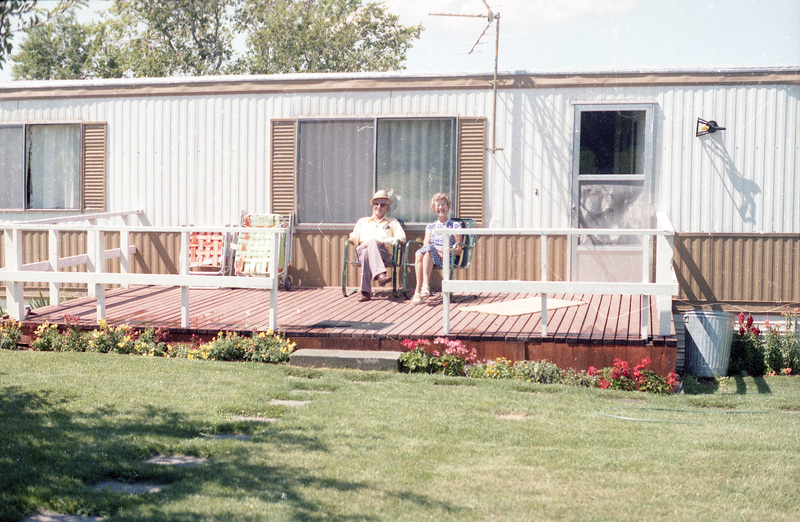 Two people sitting on chairs on a deck. There is a long white building in the background and a lawn in the foreground.