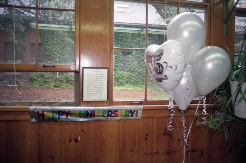 A photo of a banner that reads "Happy Anniversary!" and some balloons. The banner is hung from a wooden ledge on a wall with open windows.
