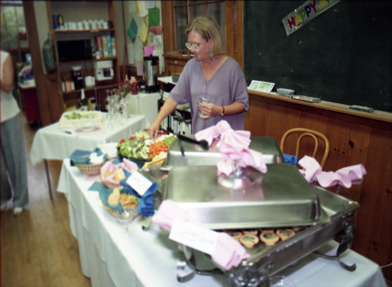 Susan Palmer grabbing food from a buffet spread. Jill Anderson can be seen off-camera to the left in a white shirt and gray slacks. There is also a blackboard with part of a banner "Happy..." visible.
