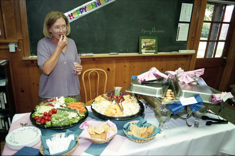 Susan Palmer eating food from a buffet spread. A part of a banner that reads "Happy Anniversary" can be seen affixed to a blackboard in the background. Another woman wearing white can be seen in the background.