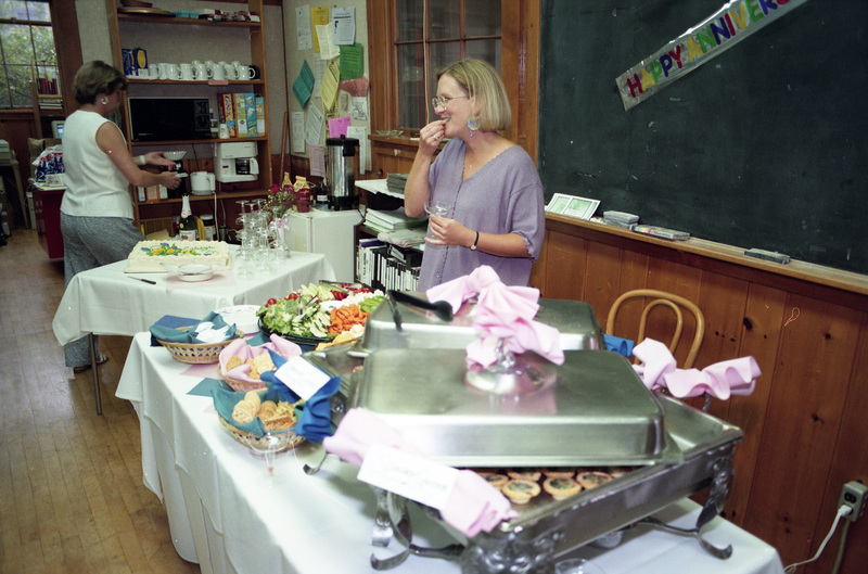 Susan Palmer eating food from a buffet spread. Jill Anderson is facing away and standing near the small kitchen area in the background. A part of a banner that reads "Happy Anniversary" can be seen affixed to a blackboard in the background.