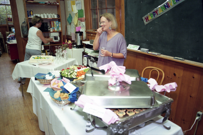 Susan Palmer eating food from a buffet spread. Jill Anderson (former Women's Center office coordinator) is in the background, near the coffee machine. A part of a banner that reads "Happy Anniversary" can be seen affixed to a blackboard in the background.