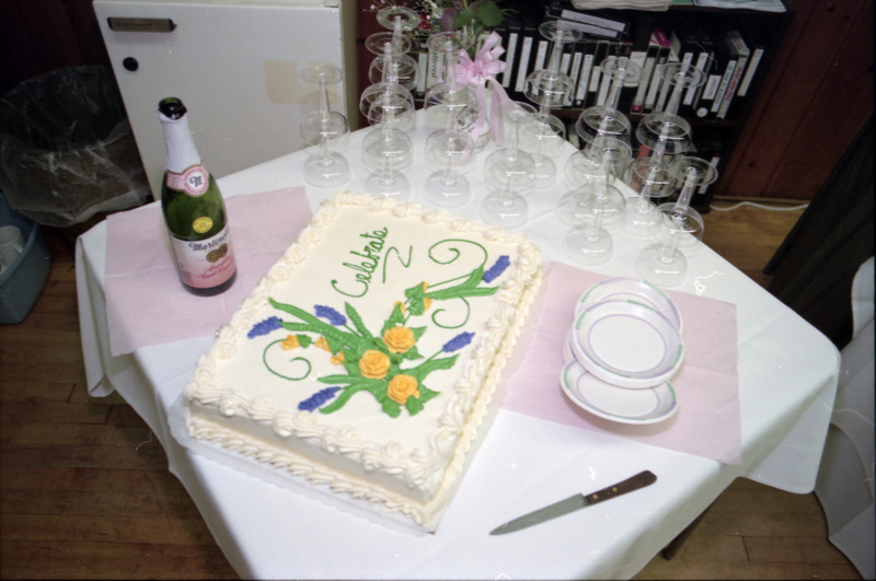 A photo of a cake which reads "Celebrate." A bottle of Martinelli's, empty glasses, and plates are also on the table. VCR tapes can be seen in the background.