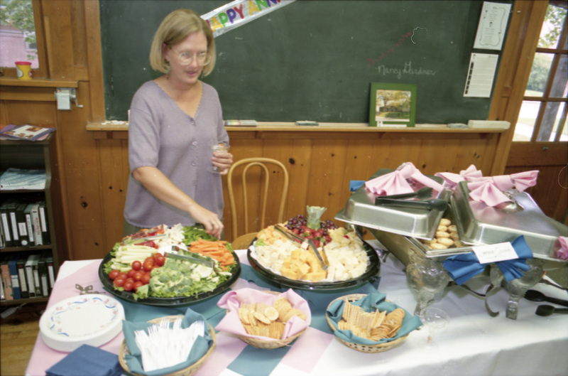 Susan Palmer grabbing a green pepper from a veggie platter on a table with a larger food spread. A banner affixed to the blackboard in the background reads, "Happy Anniversary."