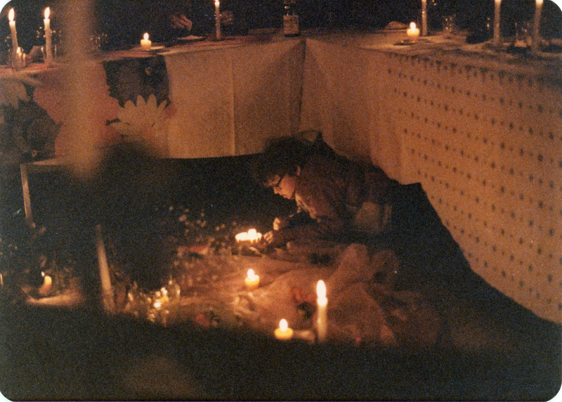 Photo from a recreation of Judy Chicago's The Dinner Party, held in St. Augie's. A person lies on their stomach tending a lit candle. They are partially underneath a table that is lined with lit candles.