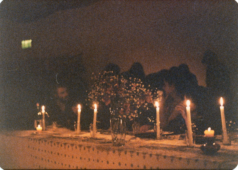 Photo from a recreation of Judy Chicago's The Dinner Party, held in St. Augie's. Several people sitting at tables. The room is lit by candlelight. Some plants can be seen in a glass on the table.