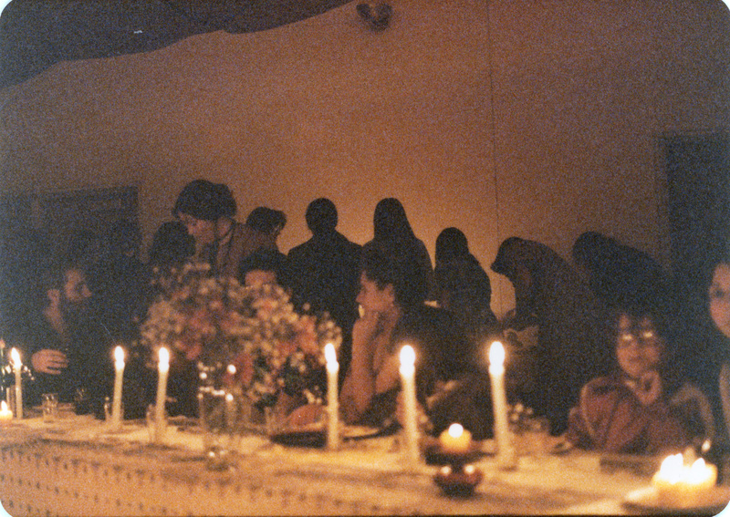 Photo from a recreation of Judy Chicago's The Dinner Party, held in St. Augie's. Several people sitting at tables. Another group of people stand in the background with their backs toward the camera. The room is lit by candlelight.