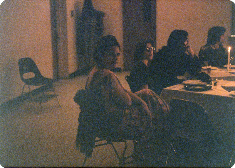 Photo from a recreation of Judy Chicago's The Dinner Party, held in St. Augie's. Several people sitting at tables including Jenifor Klindt sitting second from the left. The room is lit by candlelight.