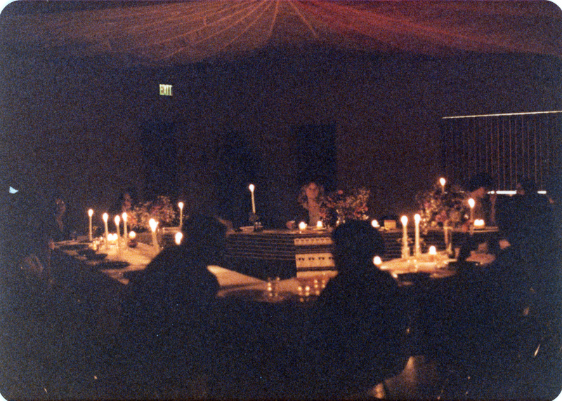 Photo from a recreation of Judy Chicago's The Dinner Party, held in St. Augie's. Several people sitting at tables facing one another. The room is lit by candlelight.