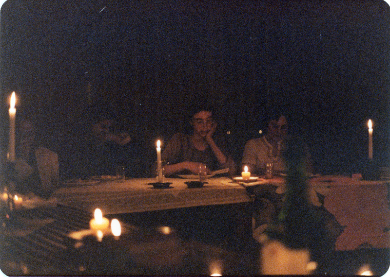 Photo from a recreation of Judy Chicago's The Dinner Party, held in St. Augie's. Several people sitting at tables. The room is lit by candlelight. Some of the people are looking down at something, possibly paper, on the table.