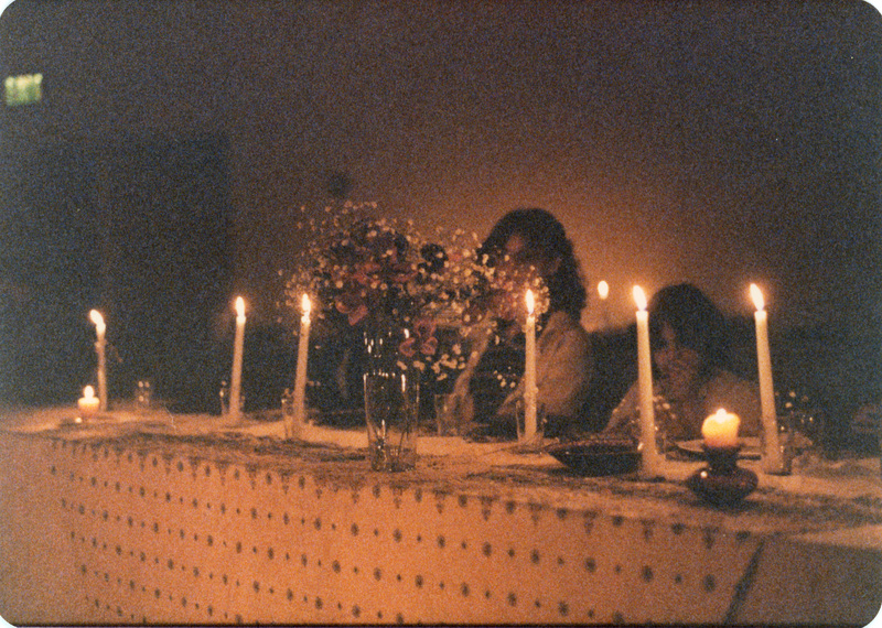 Photo from a recreation of Judy Chicago's The Dinner Party, held in St. Augie's. Several people sitting at a table. There is a clear vase with flowers on the table. The room is lit by candlelight.