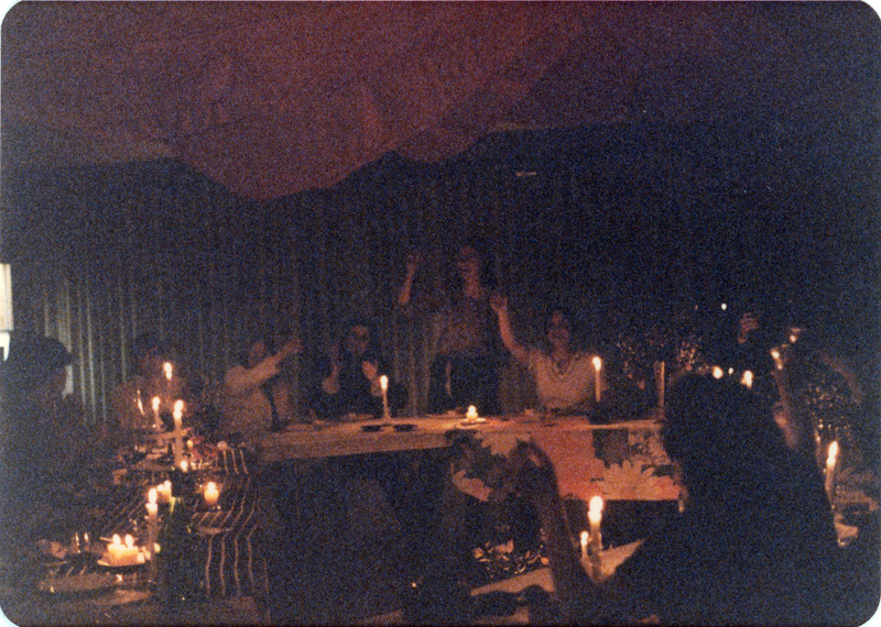 Photo from a recreation of Judy Chicago's The Dinner Party, held in St. Augie's.  Several people sitting at tables holding glasses with their arms raised. The room is lit by candlelight.
