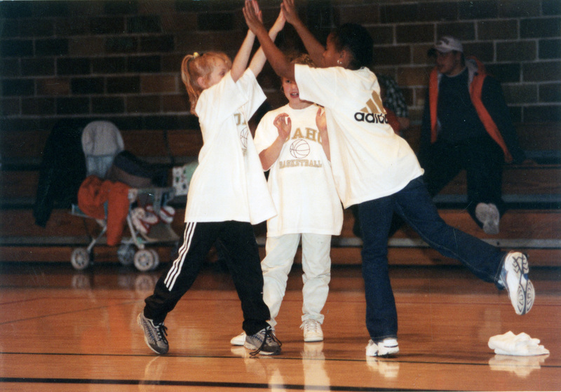 Three children on a basketball court. They are wearing shirts that read, "Idaho Basketball." Two of the children clasp hands creating a pyramidal formation while the other child stands between them.