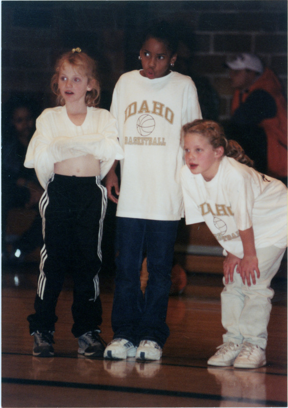 Three children on a basketball court. They wear shirts that read, "Idaho Basketball." Other people can be seen sitting on bleachers in the background.