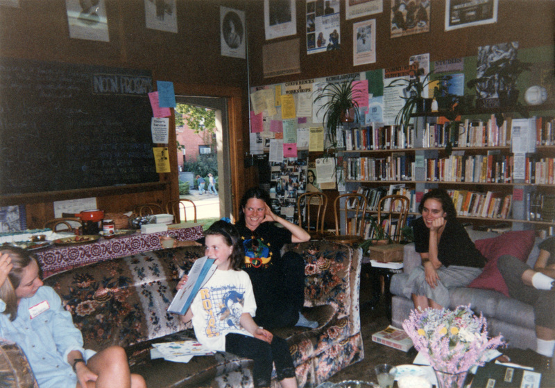 A group of three adults and one child sit on couches in the Women's Center. A blackboard, various posters and fliers, and a bookshelf can be seen in the background. 