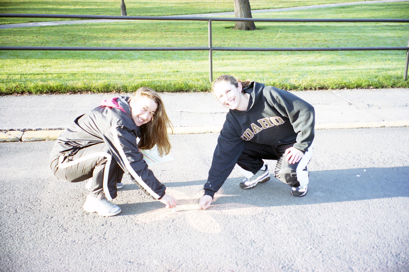 Two people drawing a flower with orange chalk on pavement.