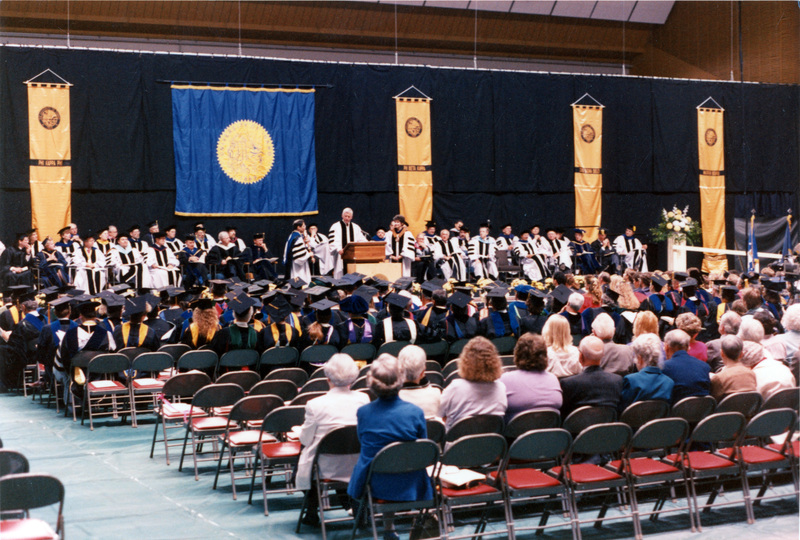 Scene during Elisabeth Zinser's inauguration. Zinser was the first female president of the University of Idaho. A man stands at the podium as people in regalia on both the stage and in the audience look on.
