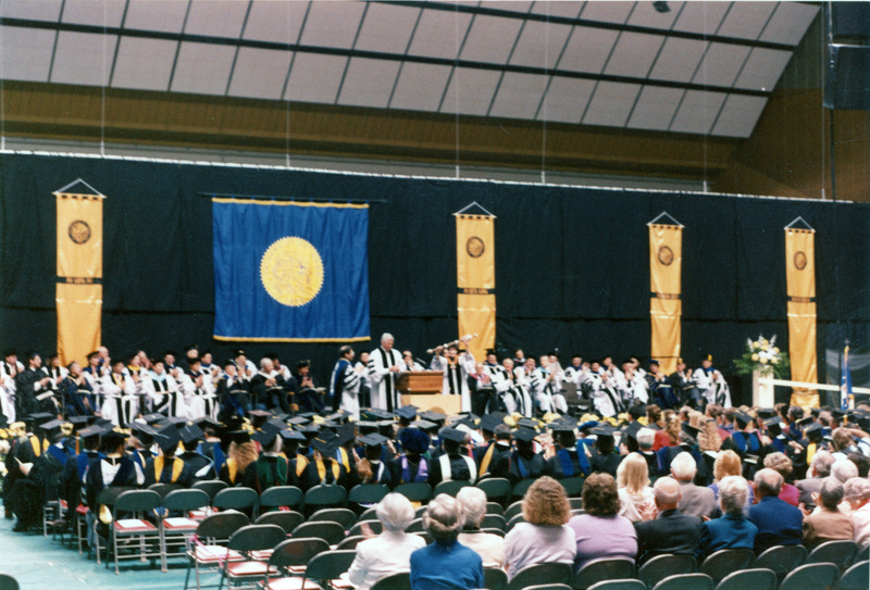 Scene during Elisabeth Zinser's inauguration. Zinser was the first female president of the University of Idaho. A man and a woman (possibly Zinser) stand at the podium as people in regalia on both the stage and in the audience look on.