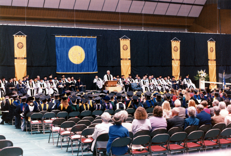 Scene during Elisabeth Zinser's inauguration. Zinser was the first female president of the University of Idaho. A man and a woman (possibly Zinser) stand at the podium as people in regalia on both the stage and in the audience look on.