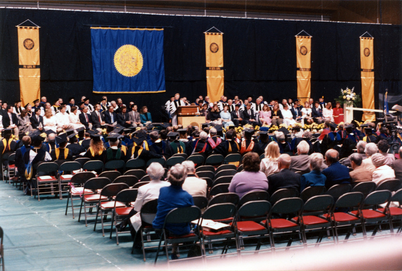 Scene during Elisabeth Zinser's inauguration. Zinser was the first female president of the University of Idaho. A woman speaks at a podium. About half of the people on stage and audience are wearing regalia.