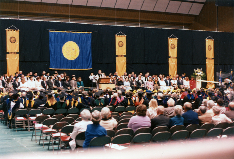Scene during Elisabeth Zinser's inauguration. Zinser was the first female president of the University of Idaho. A woman speaks at a podium. About half of the people on stage and audience are wearing regalia.