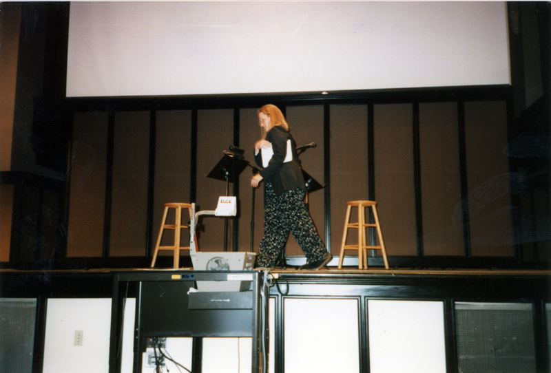 Susan Palmer on the Administration Auditorium stage. A projector is below the stage and a projector screen hangs behind Susan Palmer.