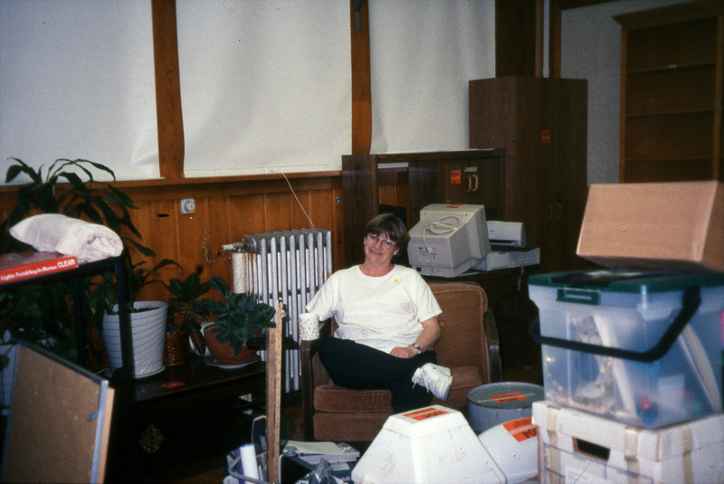 Jill Anderson sitting in a chair surrounded by boxes and office equipment. Women's Center relocation from the Old Journalism Building to the Theatre Annex, Summer 2000.