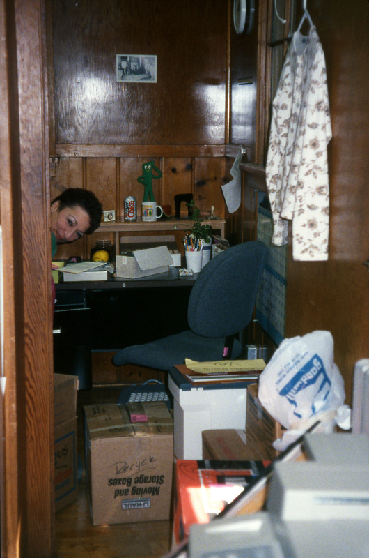 Valerie Russo peeks out from behind a door frame. There are boxes, a desk, and a chair next to her. Women's Center relocation from the Old Journalism Building to the Theatre Annex, Summer 2000.