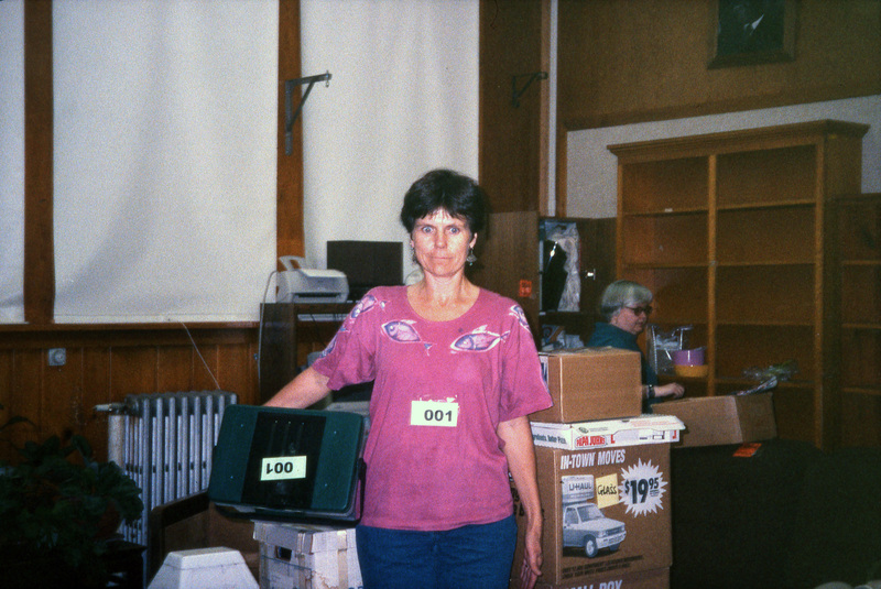 Jeannie Harvey makes a face at the camera while holding a box. There are boxes behind her and another woman in the background. Women's Center relocation from the Old Journalism Building to the Theatre Annex, Summer 2000.